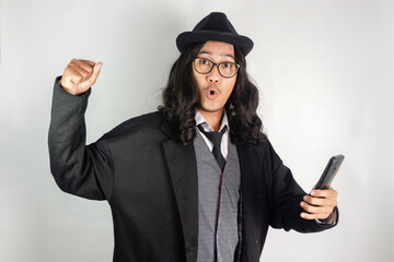 Asian man with long hair dressed as a mafia wearing glasses and hat, holding a smartphone and...