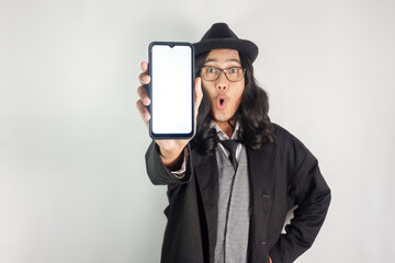 Asian man with long hair dressed as a mafia wearing glasses and a hat showing a smartphone with a...