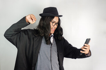 Asian man with long hair dressed as a mafia wearing glasses and a hat looking at a smartphone with...