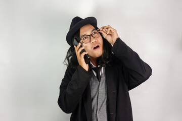 Asian man with long hair dressed as a mafia wearing glasses and a hat, making a phone call gesture with a confused look on his face