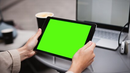 Camera focus on Caucasian girls hands. Woman holding tablet device with green screen. Tapping with finger in middle of chroma key. Sitting outside small restaurant or bar. Coffee and laptop on table.
