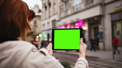 View from behind of Caucasian woman with red hair holding small tablet with both hands. Chroman key on screen of device. Green screen. Girl holding digital device steadily without moving.