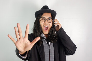 Asian man with long hair dressed as a mafia wearing glasses and a hat, making a phone call and making a refusal gesture, his face looks panicked