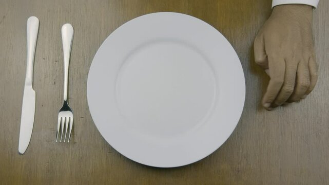 An empty plate with a fork and knife on a table, accompanied by a hand in a waiting gesture, symbolizing anticipation, readiness, and eagerness for food
