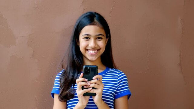 Latina teenager with cobalt blue and white striped shirt and a smartphone, smiling on a mocha mousse wall.