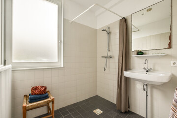 A bright, minimalist bathroom featuring simple tiles, a shower, a sink, and natural light flowing through the window, creating a serene environment.