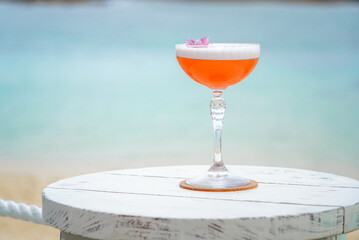 Gin sour cocktail served in an elegant coupe glass, topped with foam and garnished with a delicate pink flower. Placed on a rustic white wooden table by the beach.