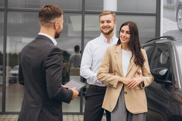 Young couple and vehicle manager standing outside auto showroom near car. Man and woman talking with sales manager.
