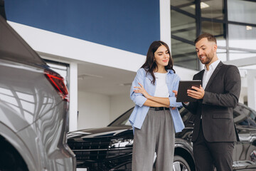 Young woman customer female buyer client wears shirt consult with salesman hold tablet choosing auto want to buy new automobile in car showroom vehicle salon dealership store motor show.