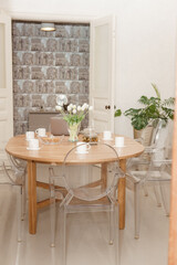 A cozy dining setup, featuring a wooden table with a laptop, tea set, and tulip centerpiece, surrounded by transparent chairs, exudes warmth and modern aesthetics.