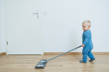 Fototapeta premium Blond toddler boy washes the floor with a mop, helping his mother clean at home, free space. Child learns to clean the house.