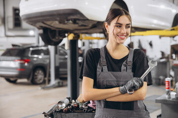 Smiling young woman mechanic standing beneath lifted car, holding tools and looking at camera in...