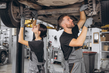 Two focused professional mechanics, bearded man and young woman, wearing grey overalls, repairing undercarriage of car on lift in auto repair shop