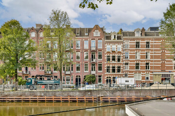 Naklejka premium Picturesque row of historic buildings along a serene canal, exemplifying Amsterdam's unique architectural style with vivid facades and lush greenery.