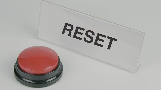 "Reset" text or word displayed on a nameplate as a hand presses a red button, set against a white background, symbolizing the act of restoring settings, starting over, or making a fresh start