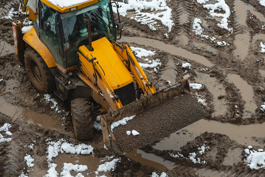 A loader tractor with a bucket filled with gravel is driving through muddy and wet off-road at a construction site. Construction is taking place during the winter season.
