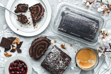 Delicious chocolate brownie roll cake stuffed with cream, cherries and nuts on light background with flowers and cup of coffee. Sweets, dessert and pastry, top view