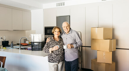 Smiling elderly couple embracing and drinking coffee in their new home kitchen with cardboard boxes...