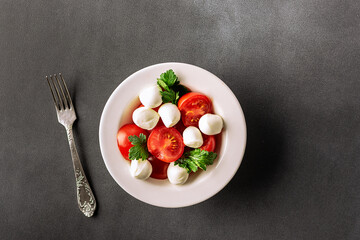 Juicy caprese salad with baby mozzarella cheese, cherry tomatoes and parsley in white ceramic plate on black stone table top view. Healthy diet food concept.