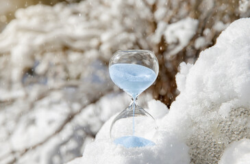Conceptual image of switching to winter time or fall back, end of daylight saving time. Selective focus on hourglass with blue color sand falling in the winter in snowy nature.