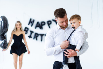 Dad and son at birthday party. A father holds his son in formal attire as a woman in black decorates for a birthday behind them.