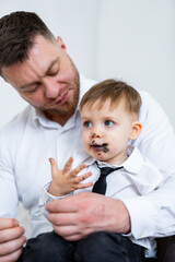 Father and son enjoy dessert together. A father sits with his young son, who has chocolate smudges...