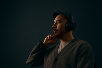 Portrait of a handsome caucasian man with a beard sitting in a chair in a dark grey room wearing a cardigan and wireless black headphones listening.