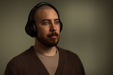 Portrait of a handsome caucasian man with a beard and a shaved head standing in a studio wearing a cardigan and wireless black headphones listening.