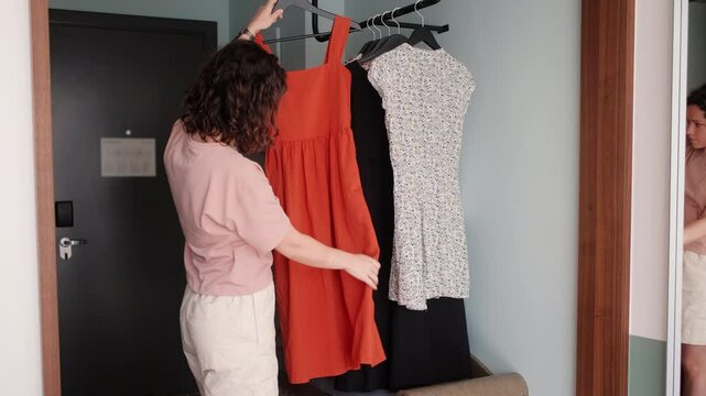 woman standing in hotel room choosing between dresses, holding outfit in front of mirror, planning stylish look for the day, travel morning routine, fashion decision in cozy modern interior