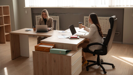 The image shows two professional women working in a modern office. One is typing on a laptop, while the other is enjoying a coffee, with documents and laptops on the shared desk.
