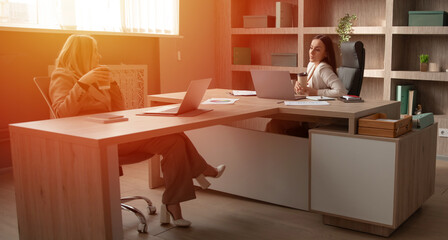 Obraz premium Two professional women in a bright office. One is sitting and holding a coffee, while the other is working on a laptop. The space features modern furniture, a shared desk, and organized shelves.