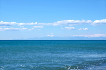 Beautiful panoramic landscape with high mountains silhouettes with snow cap on tops far on skyline in light haze behind blue sea under clear sky with some white clouds