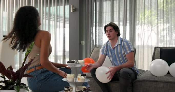 Happy diverse couple blowing up balloons and preparing for birthday party in living room