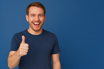 Smiling young man in a navy blue t-shirt giving a thumbs up against a solid blue background