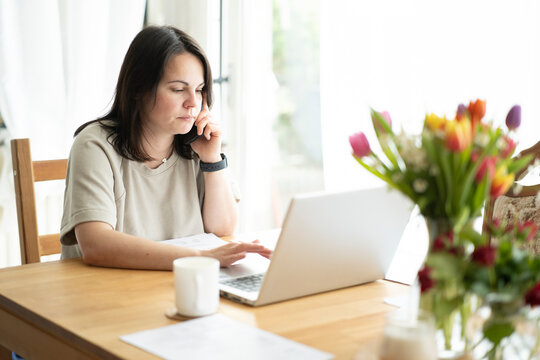Beautiful dark-haired girl talking seriously on the phone sitting at a wooden table in front of a laptop and documents, a mug of coffee on the table. Home office concept