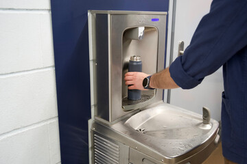 A traveler uses a stainless steel fountain to refill a dark reusable bottle inside an airport, showcasing a sustainable water source next to a door and blue panel