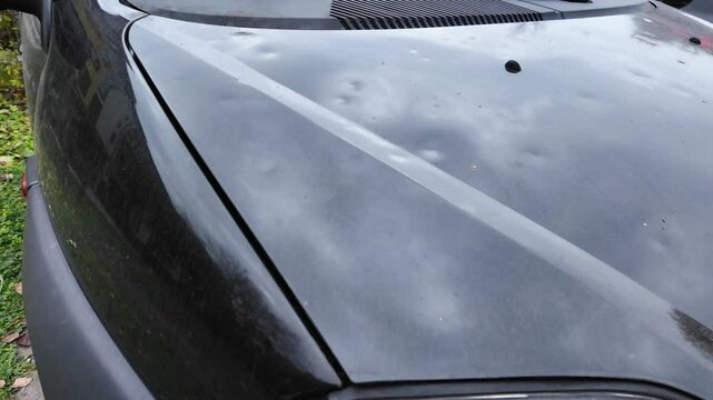 Hood of a black car damaged by hail storm. Dents caused by ice grains. Parking outdoors in extreme weather conditions. Everyday problems, car insurance concept.