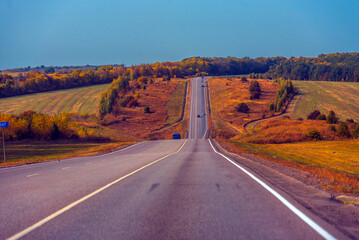 A long, straight paved road leads into the distance through a hilly autumn landscape. The sides of the road and the surrounding fields are painted in the golden-red tones typical of autumn 