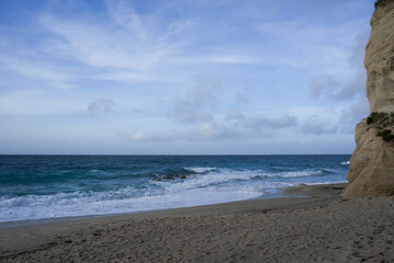 A peaceful sandy beach is bordered by rugged cliffs and touched by gentle blue ocean waves beneath a soft sky with wispy clouds near the waterline