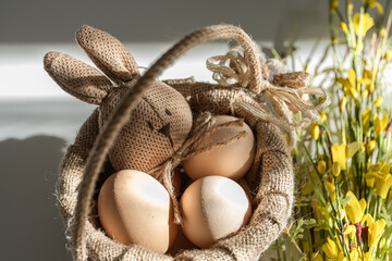 A rustic Easter-themed still life featuring a woven basket filled with natural brown and white eggs, along with a handmade burlap bunny character. 