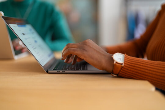 A close-up view of a person's hands typing on a laptop keyboard, with a focus on their wristwatch and a blurred office background featuring another person working.