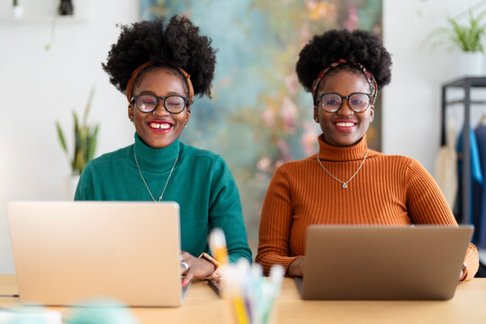 Two young women with curly hair, glasses, and stylish outfits are smiling while working on laptops at a wooden table in a bright and modern coworking space.