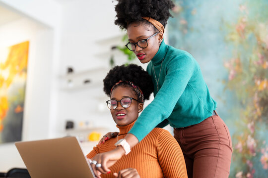A woman in a green sweater leans over to help her colleague with a laptop in a bright office with modern decor, featuring indoor plants and a clothing rack.