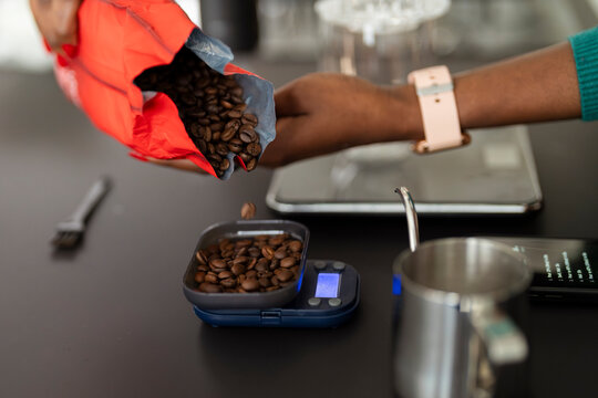A close-up of a person pouring coffee beans from a red bag onto a digital scale, preparing to measure the precise amount for brewing, with a coffee-making setup in the background.
