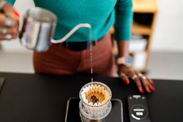 A woman pours hot water from a sleek metal kettle into a glass pour-over dripper, slowly wetting the coffee grounds in a careful brewing process in a modern kitchen setting.
