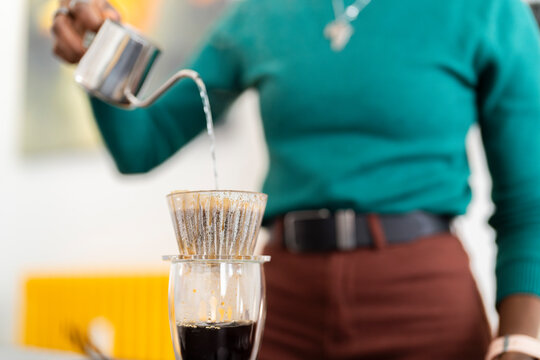 A close-up shot of a steady stream of hot water being poured from a metal kettle into a glass pour-over dripper, highlighting the slow and careful brewing process in a kitchen.