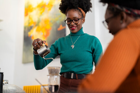 A smiling woman in a green sweater and glasses pours hot water from a metal kettle into a pour-over coffee dripper while engaging in a pleasant conversation in a cozy kitchen.