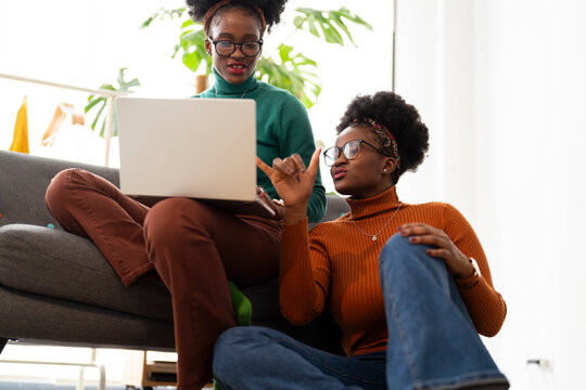 Two women sit on a gray couch, one holding a laptop while the other looks on with a smartphone, engaging in a friendly and focused moment in a bright home setting.