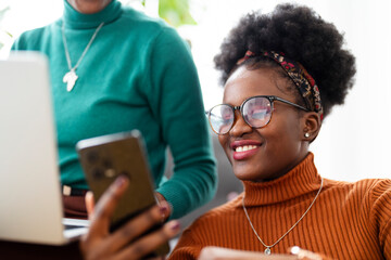 A woman holding a smartphone smiles while looking at her friend who is using a laptop, both enjoying a friendly and connected moment in a bright, cozy home setting.