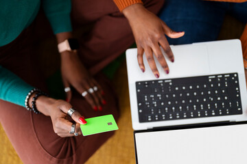 Two women sit on the floor with a laptop, one entering details while the other holds a credit card, making an online purchase in a bright, modern home setting.